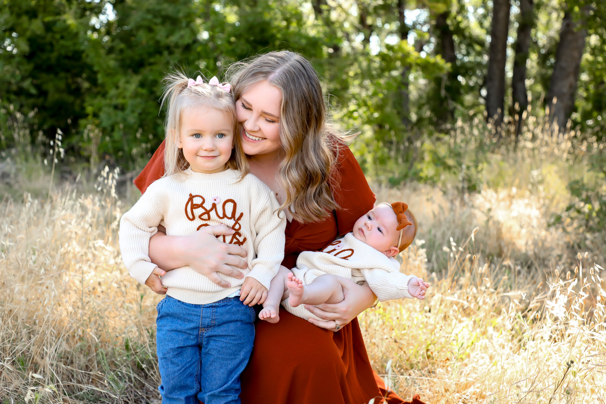 A woman in a rust-colored dress kneels in a grassy field, smiling at a young girl in a Big Sis sweater. She holds a baby in a matching Little Sis sweater. Trees and sunlight fill the background.