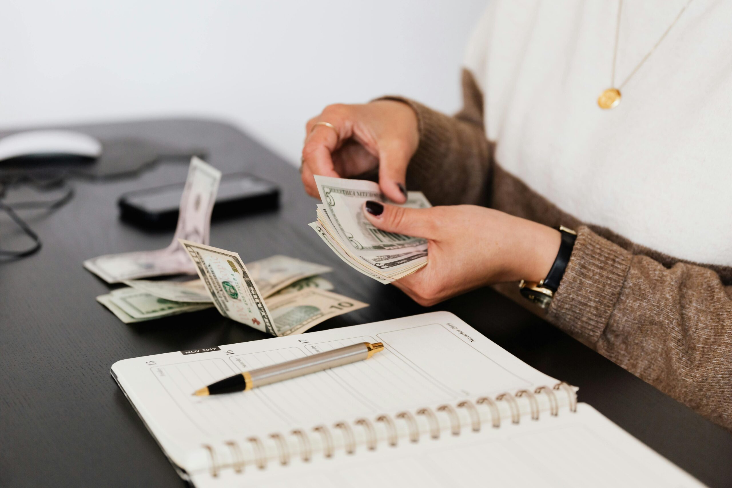 A person is counting U.S. dollar bills at a desk with a notebook, pen, and scattered cash—reminding small business owners to stay alert for QuickBooks Online scams while managing their finances. A phone and computer mouse are also visible in the background.