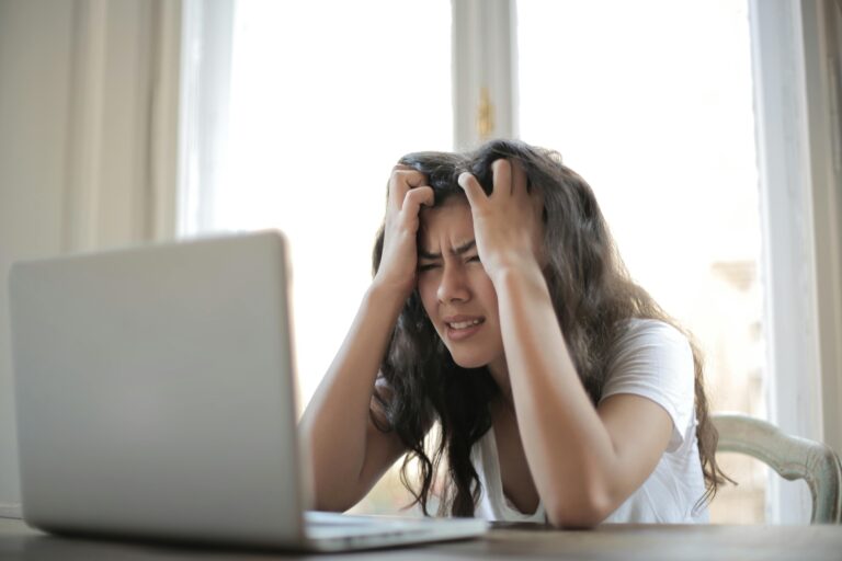 A woman sits at a desk in front of a laptop, holding her head with both hands and appearing frustrated or stressed—perhaps overwhelmed by tracking her financial habits. Light streams in through a window behind her.
