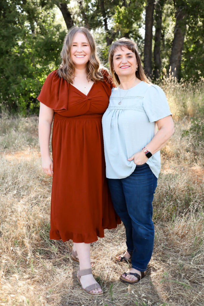 Two women stand side by side outdoors on dry grass, smiling at the camera. One wears a rust-colored dress and sandals; the other wears jeans, a light blue top, and sandals. Trees and greenery are in the background.