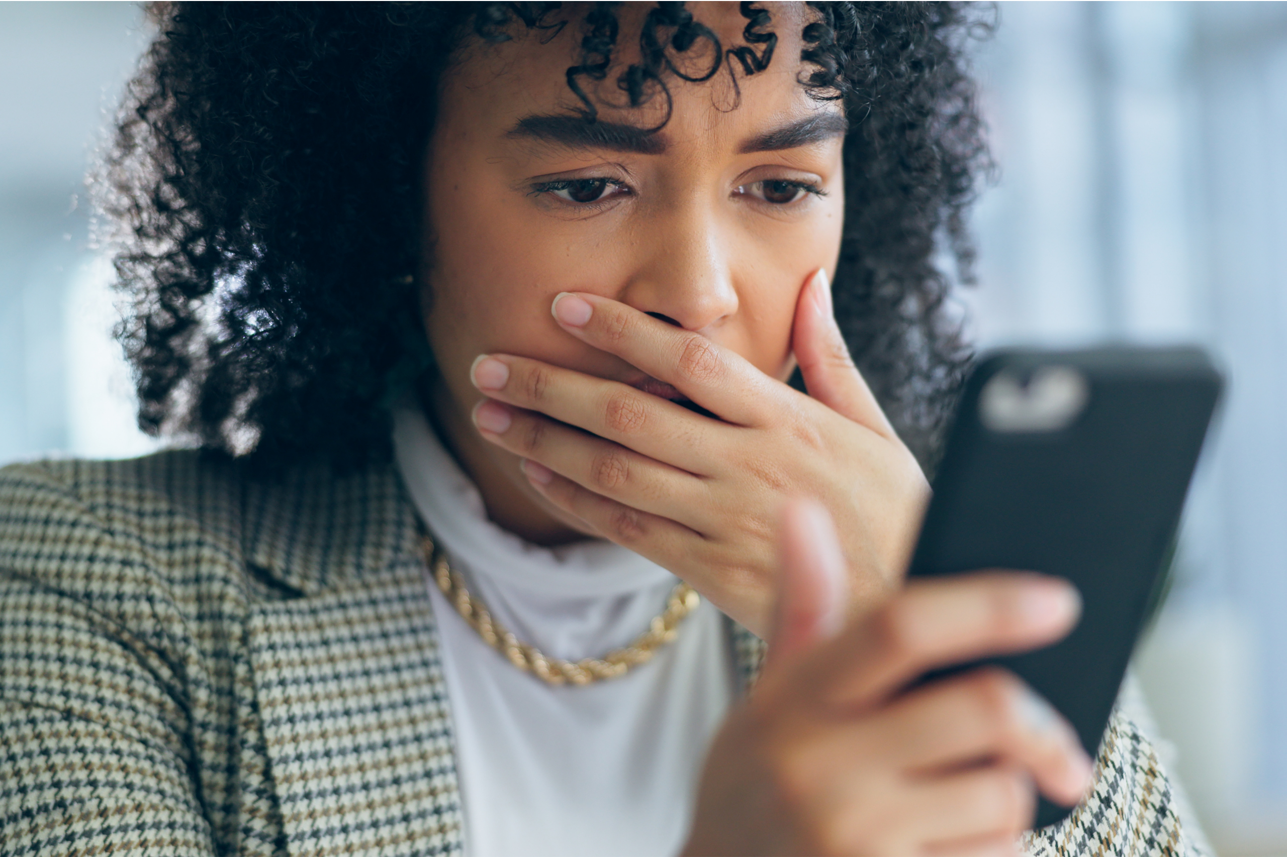 A person with curly hair wearing a blazer and gold chain looks at their phone with a worried expression, covering their mouth with one hand—possibly reacting to a QuickBooks invoice scam.