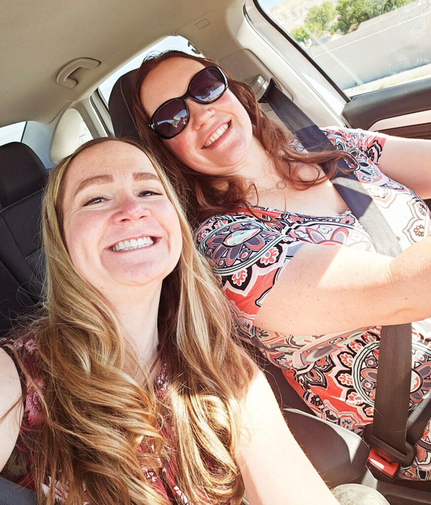 Two women smiling and taking a selfie inside a car. The passenger is holding the camera, and the driver is wearing sunglasses and a patterned shirt. Sunlight is shining through the windows and both appear happy.