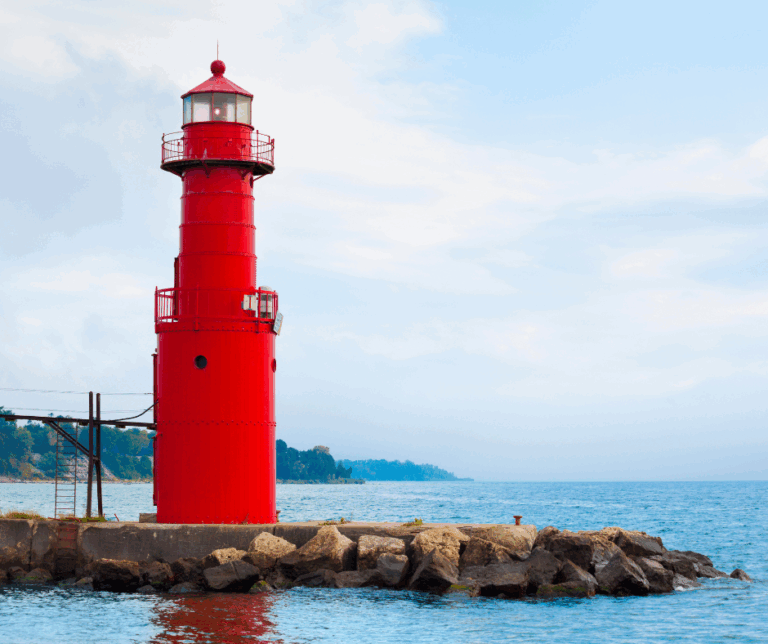 A bright red lighthouse stands on a rocky pier by the blue water, steady as small business profit—visible through clear bookkeeping, even amid clouds and distant shoreline in the background.