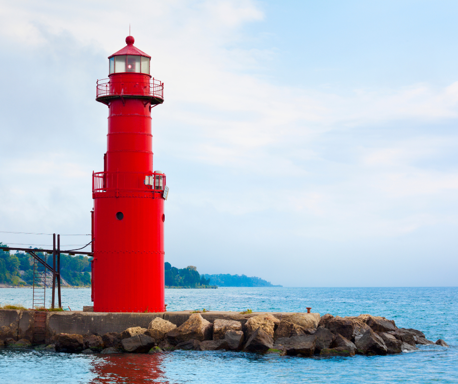 A bright red lighthouse stands on a rocky pier by the blue water, steady as small business profit—visible through clear bookkeeping, even amid clouds and distant shoreline in the background.
