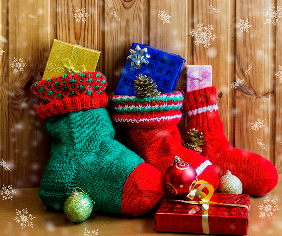 Three colorful Christmas stockings filled with wrapped gifts hang in front of a wooden wall, decorated with ornaments and snowflakes, with presents and baubles placed nearby on the floor.
