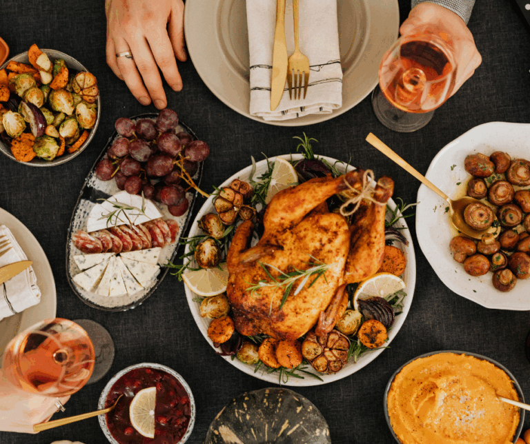 A table set with roasted chicken, potatoes, Brussels sprouts, mushrooms, cheese, grapes, hummus, cranberry sauce, and wine. Two people are holding glasses, and their hands rest near plates and napkins.