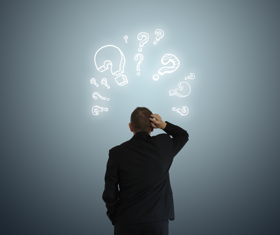 A man in a black suit stands with his back to the camera, scratching his head while looking at several white question marks drawn in the air on a gray gradient background, suggesting confusion or uncertainty.