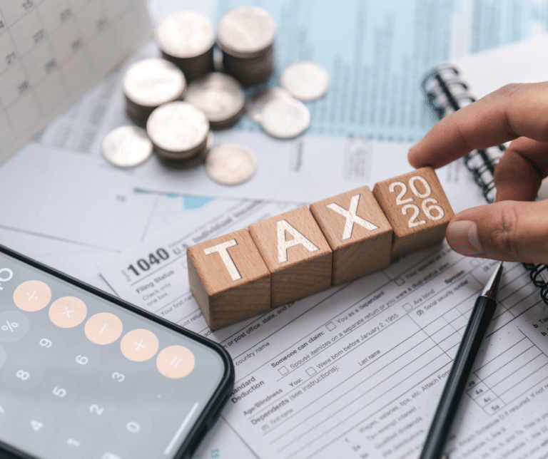 Close-up of wooden blocks spelling TAX 2026 on tax forms, with coins, a calculator, a pen, and a spiral notebook in the background, representing the concept of taxes for the year 2026.
