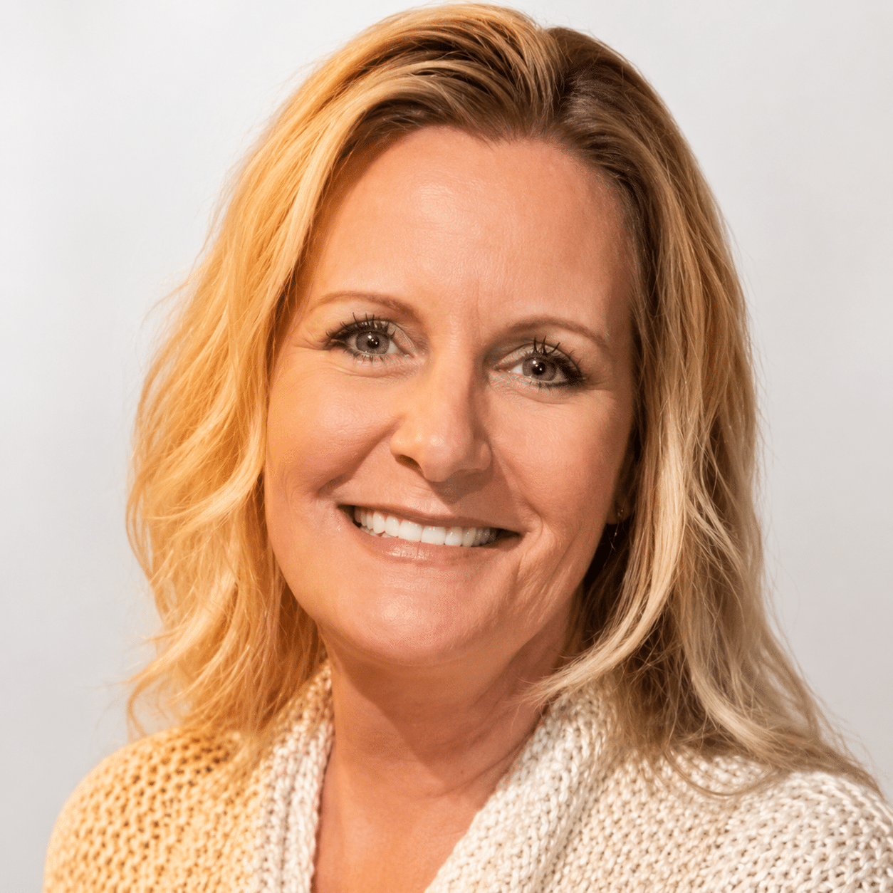 A smiling middle-aged woman with blonde, shoulder-length hair and blue eyes, wearing a cream-colored knit sweater, poses in front of a plain light background.